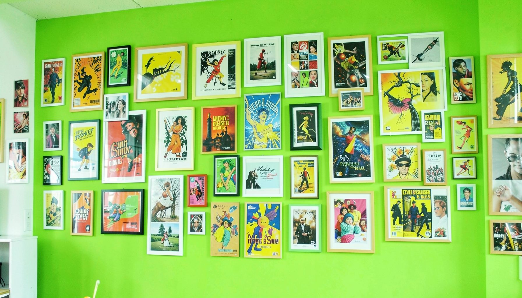 A wide collage-style wall filled with colorful framed and unframed poster art in a bright room, dominant grass green and charcoal tones creating a lively gallery mood