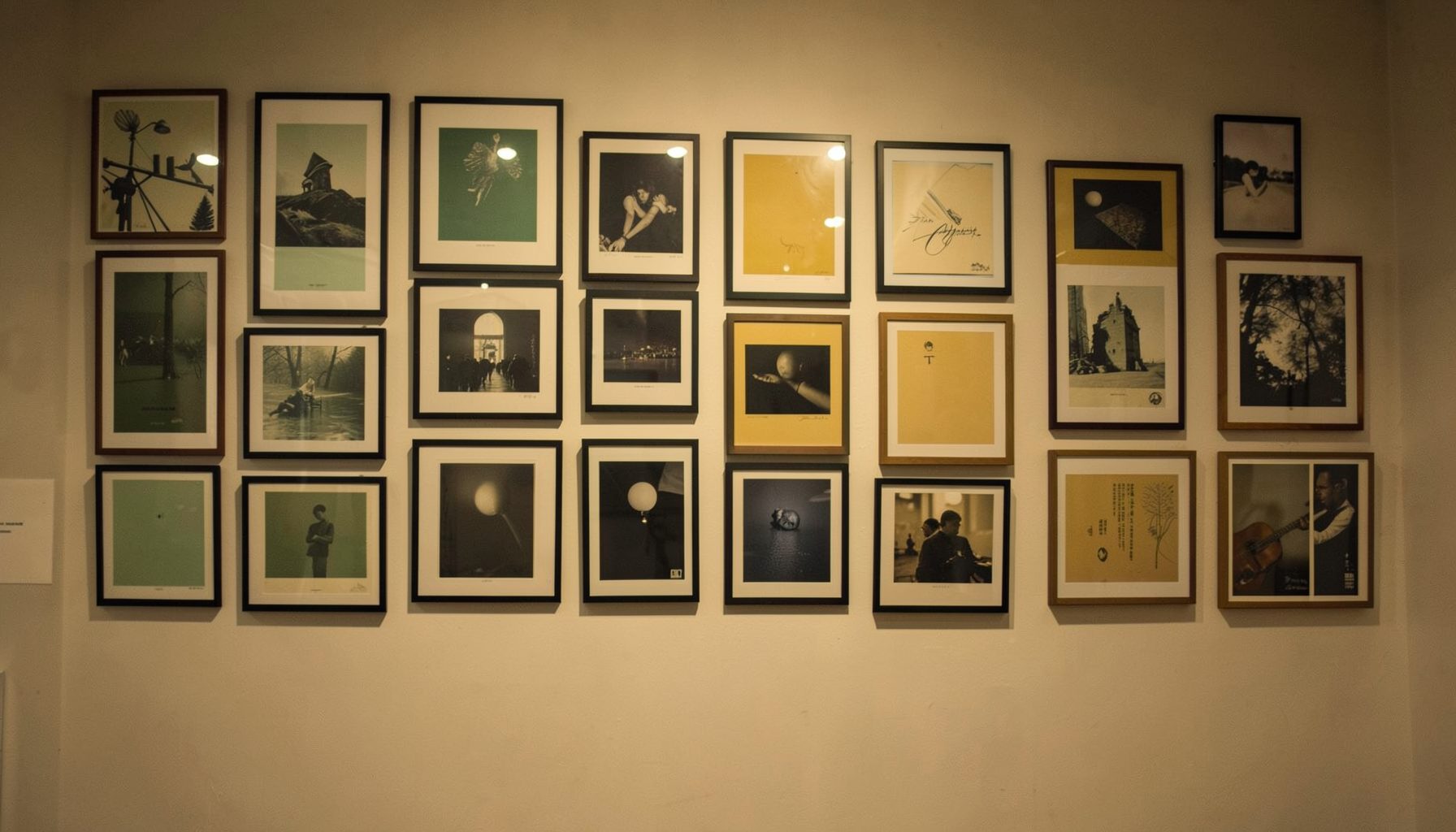 A warm, softly lit gallery wall covered with framed and unframed posters in muted green, charcoal, and mustard yellow tones